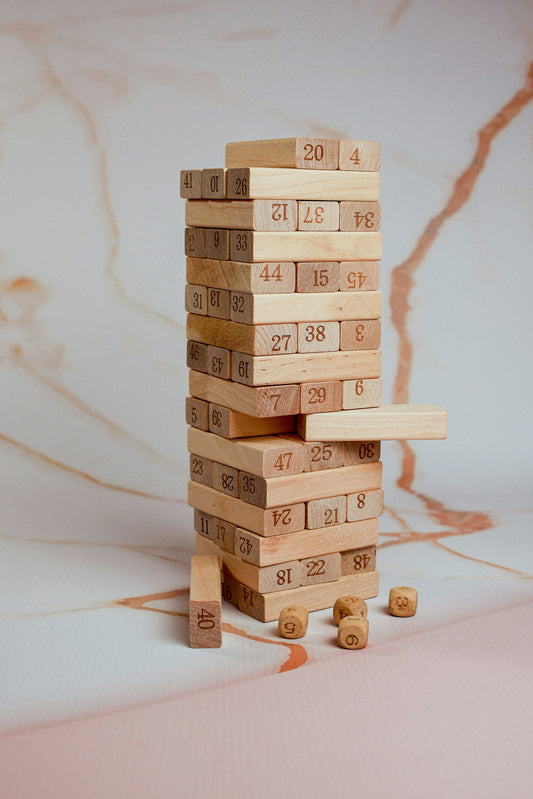 Recommended as an infographic image: a clean branded graphic showing (1) How to play in 3 steps — Build · Roll · Pull, (2) Skills developed — Numbers, Motor Skills, Strategy, Focus, (3) Who it's for — Ages 3 to adult. Alternatively, use a fun shot of the tower mid-collapse with a laughing player. The infographic version performs better on Amazon and Flipkart as it replaces the need to read bullet points for scanning buyers.